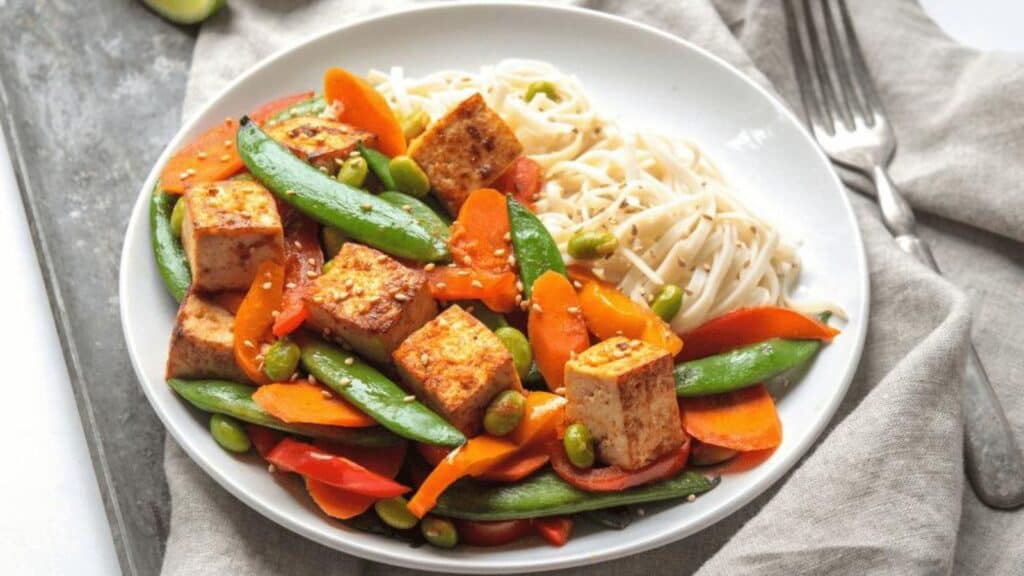 A plate of stir-fried tofu with snap peas, carrots, and noodles, seasoned with a spicy sauce.