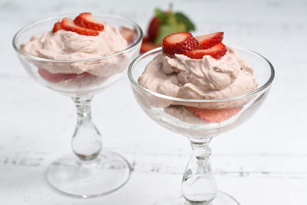 Two glass bowls of strawberry mousse topped with fresh strawberry slices on a white wooden table.