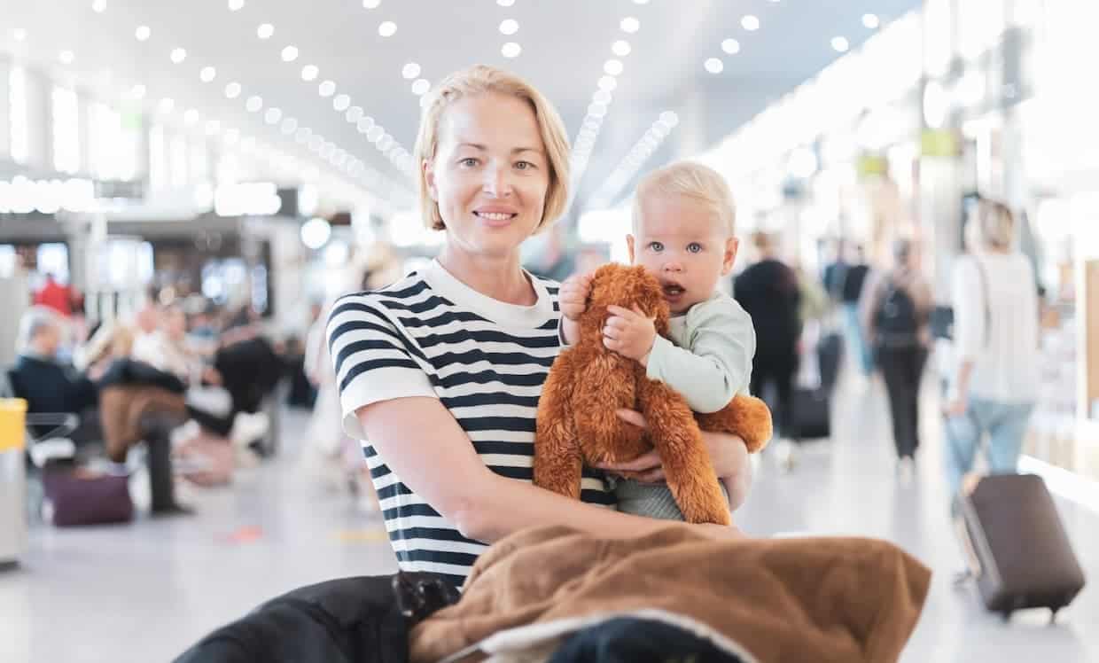 Woman holding a toddler with a stuffed toy at an airport terminal.