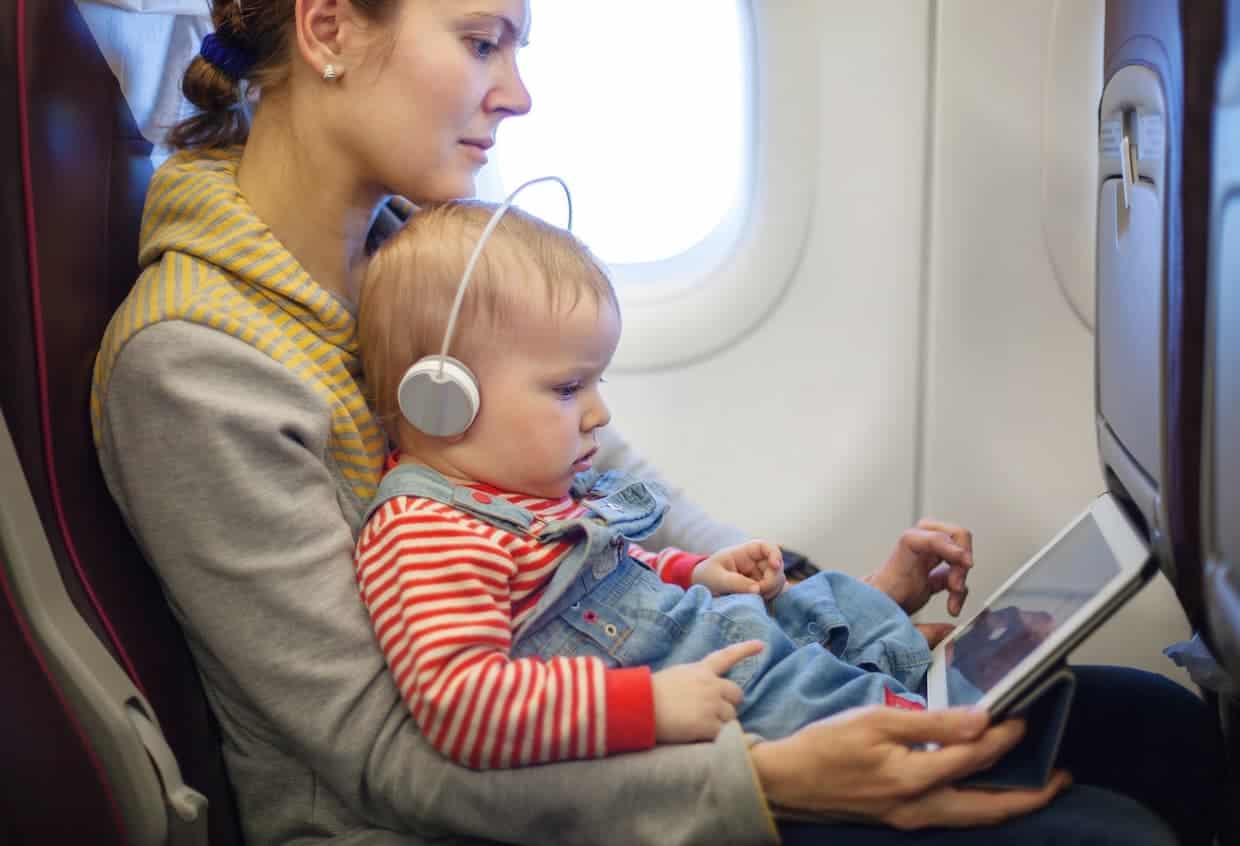 A mother and her young child wearing headphones while watching a tablet on an airplane.
