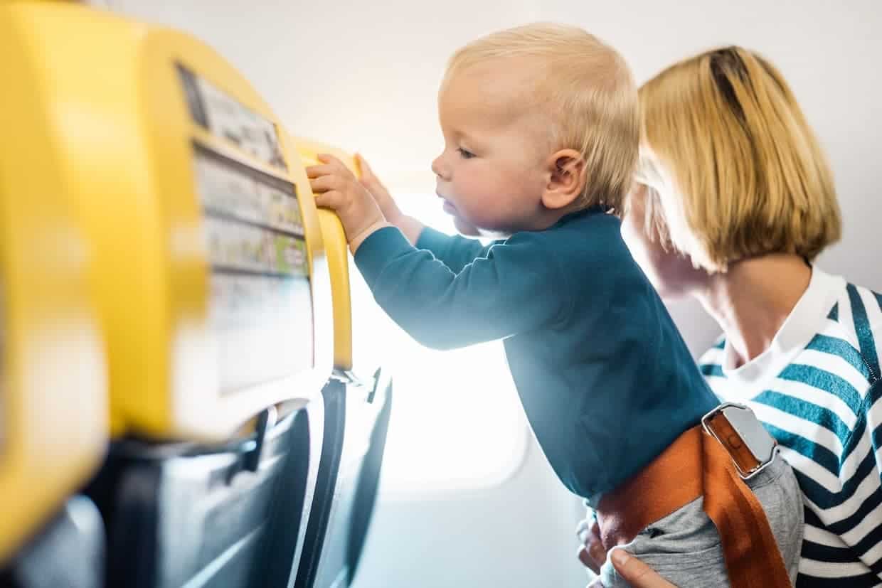 Toddler standing on an airplane seat with his mom holding him beside him.