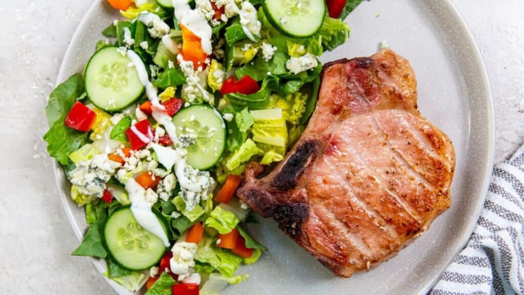 A smoked pork chop next to a fresh vegetable salad with cucumbers, tomatoes, lettuce, and blue cheese on a plate.