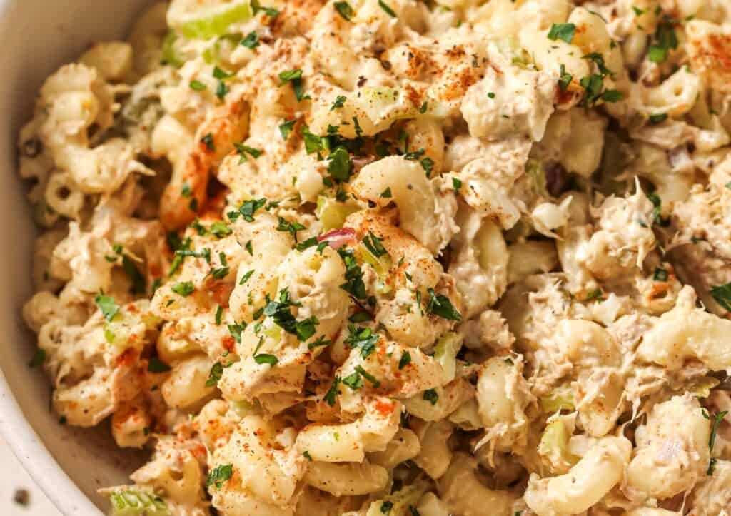 Close-up of a creamy macaroni salad with tuna and a sprinkle of paprika and chopped parsley in a white bowl.