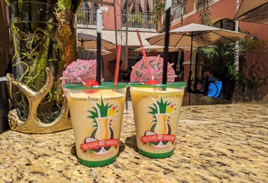 Two tropical cocktails in souvenir cups on an outdoor table, with colorful buildings and a patio umbrella in the background.