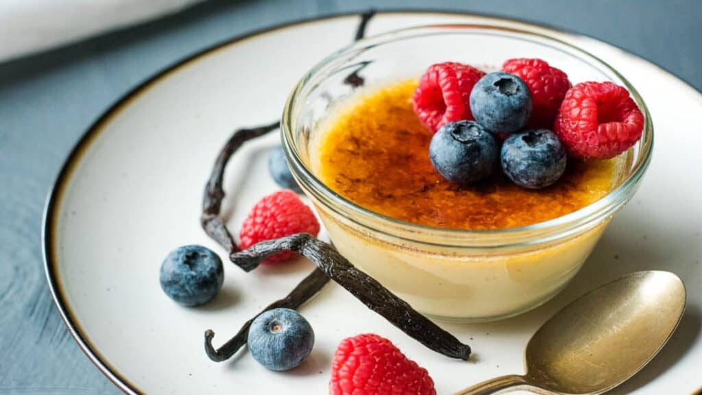 A bowl of crème brûlée topped with raspberries and blueberries beside vanilla pods.