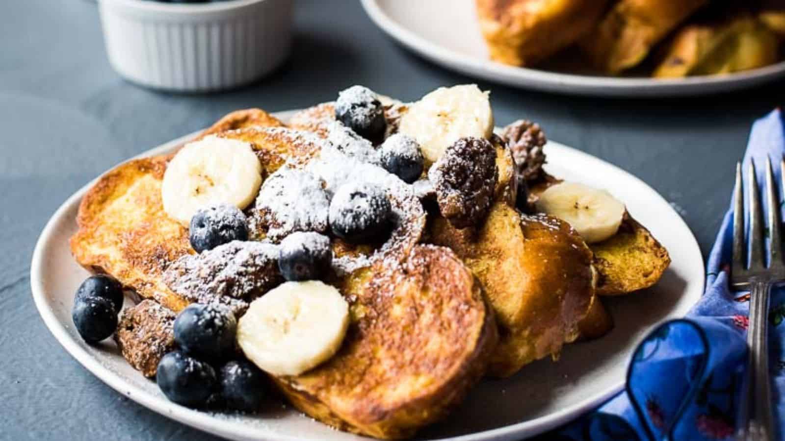 A serving of vanilla french toast next to a serving plate and bowls of candied pecans, blueberries, powdered sugar, and a banana.