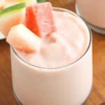 A creamy pink smoothie garnished with watermelon and melon pieces, served in a glass on a wooden table.
