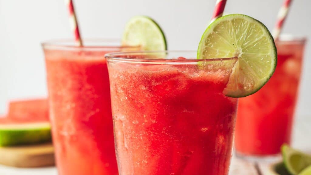 Three glasses of watermelon slushie with lime slices and red straws, set against a white background, offering sweet sips without the guilt.