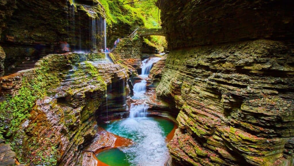 A scenic view of a lush, narrow gorge with layers of rock and cascading waterfalls surrounded by vibrant green foliage.