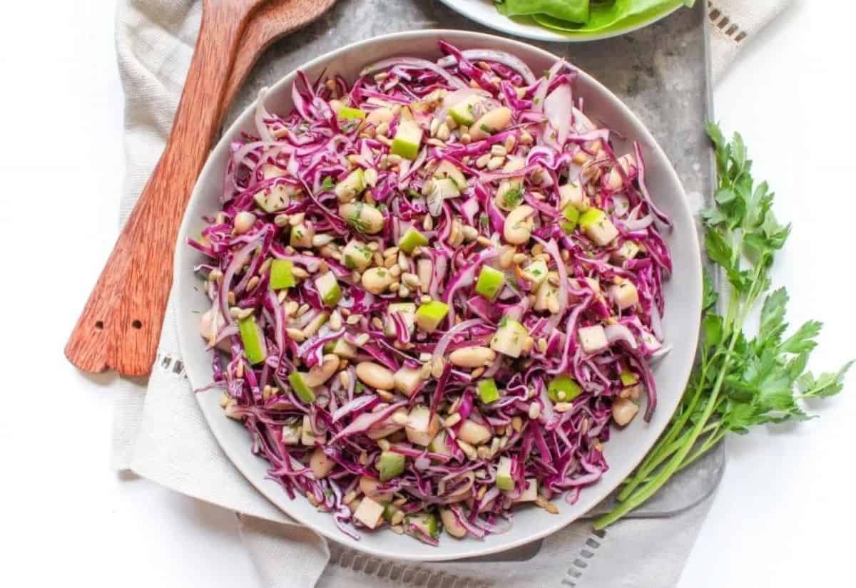 A white plate with cabbage slaw, sliced apples, and white beans on a metal sheet pan with a beige linen napkin.