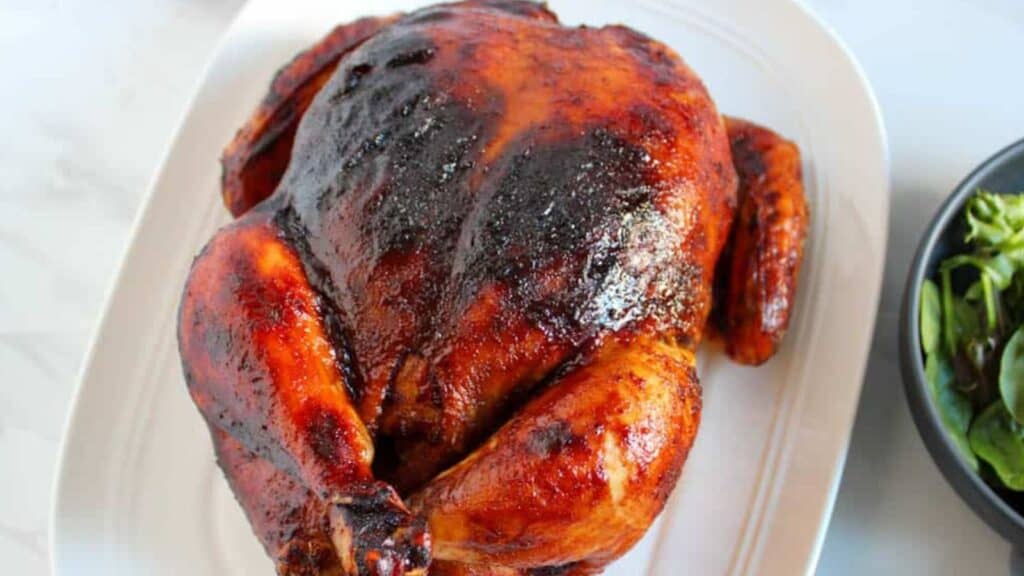 Roasted chicken with a crispy, golden-brown skin served on a white oval plate, accompanied by a bowl of green salad.