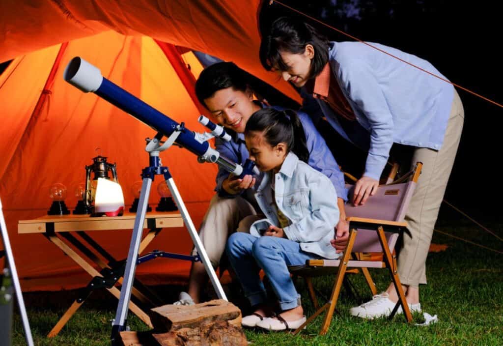 A family of three uses a blue telescope borrowed from the public library outside a lit orange tent at night. The child looks through the telescope while the adults stand nearby, smiling.