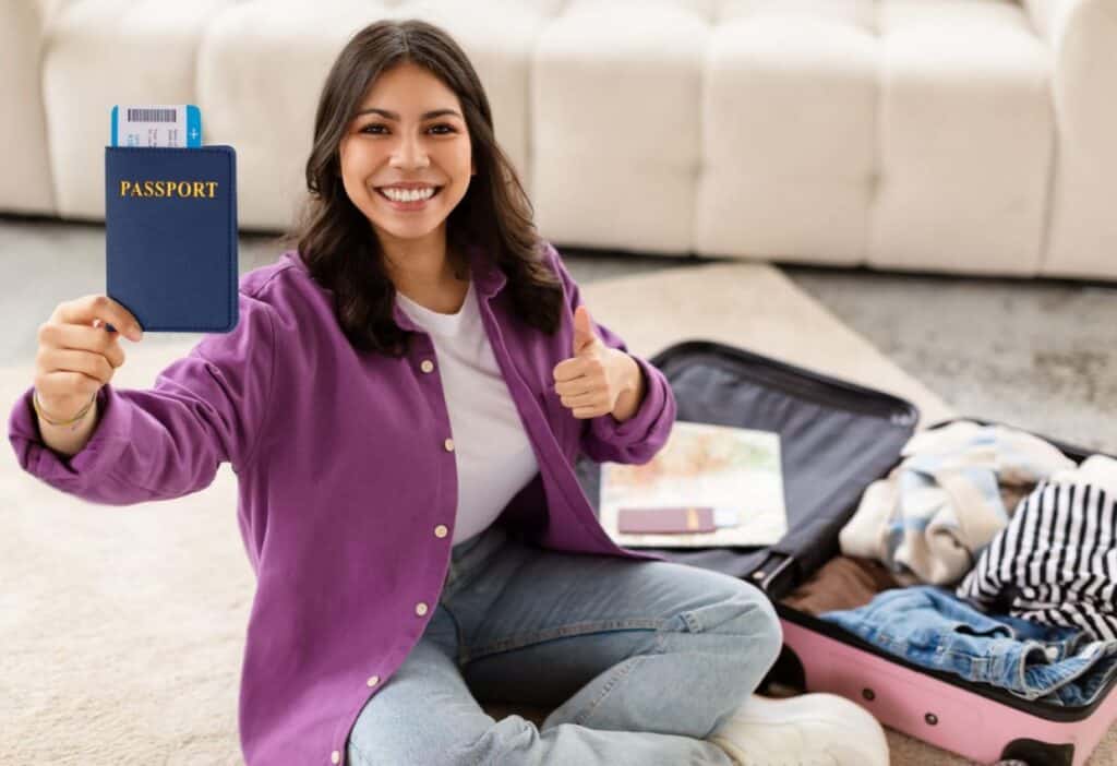 Happy woman holding a passport and giving a thumbs up while sitting with an open suitcase, ready to travel.