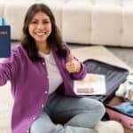 Happy woman holding a passport and giving a thumbs up while sitting with an open suitcase, ready to travel.