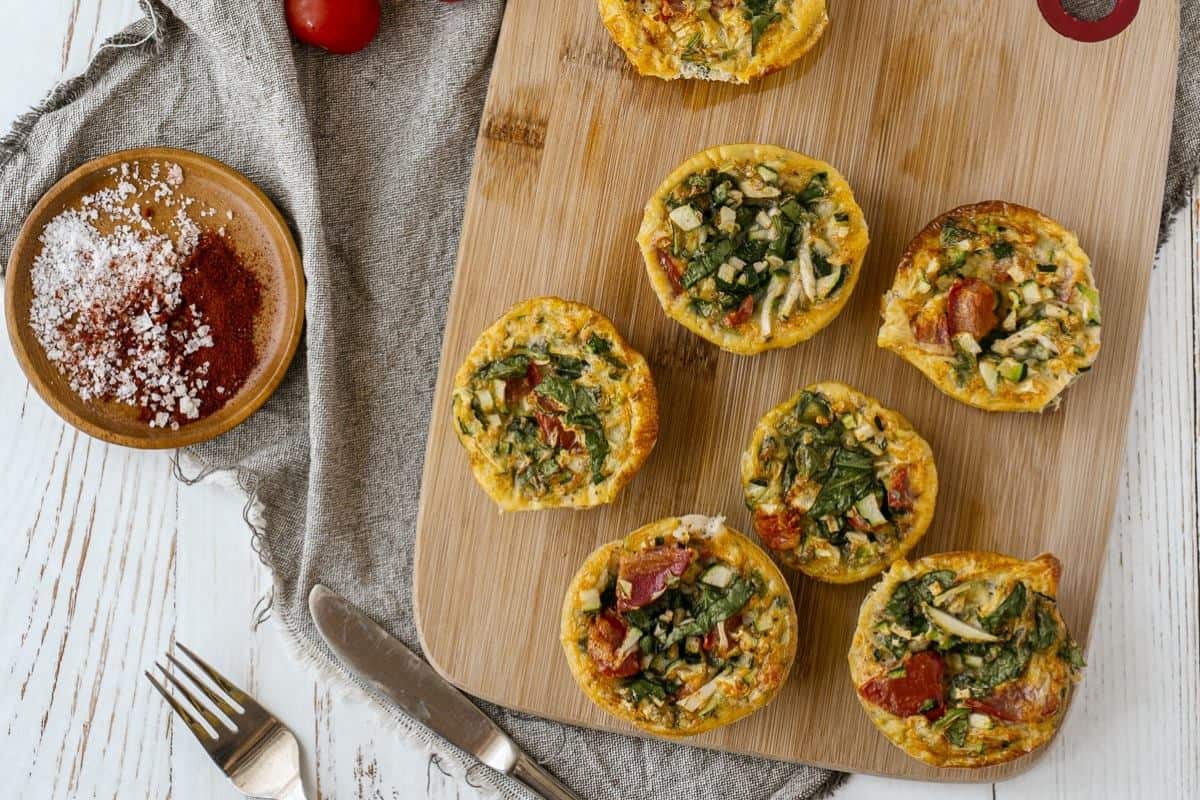 Assorted mini quiches on a wooden cutting board with a knife and seasonings to the side.