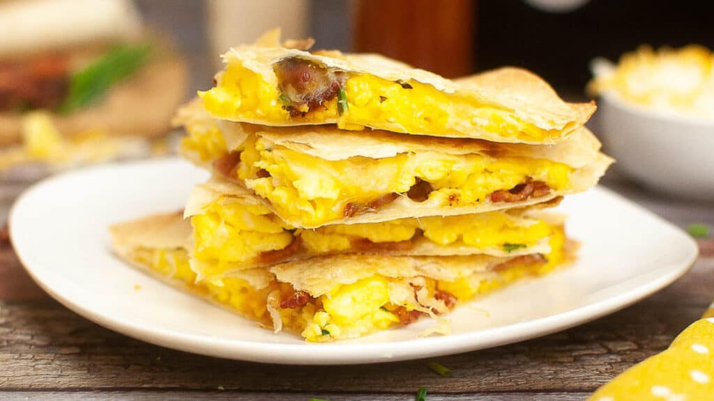 A stack of breakfast quesadillas with scrambled eggs, cheese, and bacon on a white plate.