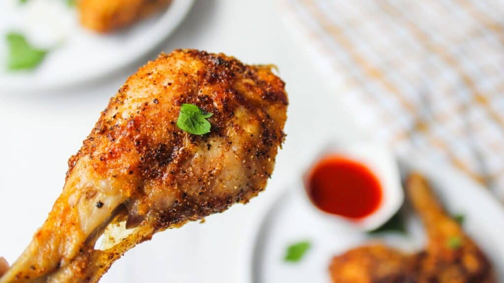 Close-up of a crispy air fried chicken drumstick on a white plate garnished with parsley, with hot sauce in the background.