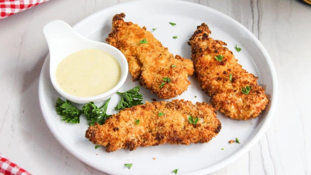 Crispy breaded chicken tenders served on a white plate with a side of honey mustard sauce and garnished with parsley.