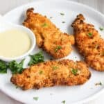 Crispy breaded chicken tenders served on a white plate with a side of honey mustard sauce and garnished with parsley.