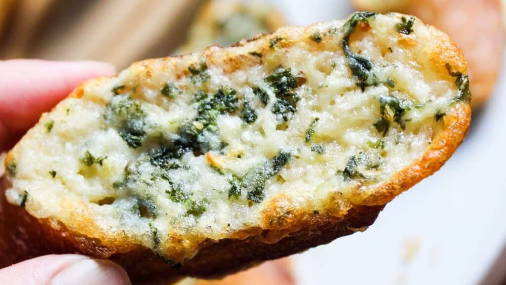 Close-up of a garlic bread slice topped with melted cheese and herbs held over a plate.