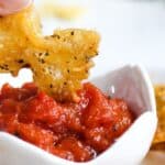 A person dips a crispy pasta chip into a bowl of marinara, with more chips visible in the background.