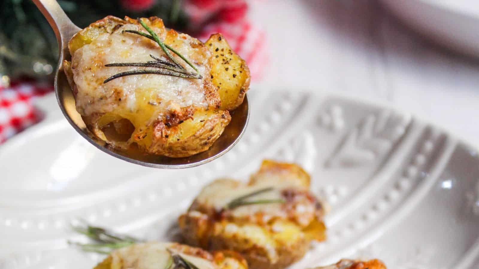 A spoon lifting a smashed potato garnished with rosemary, with a festive table setting in the background.