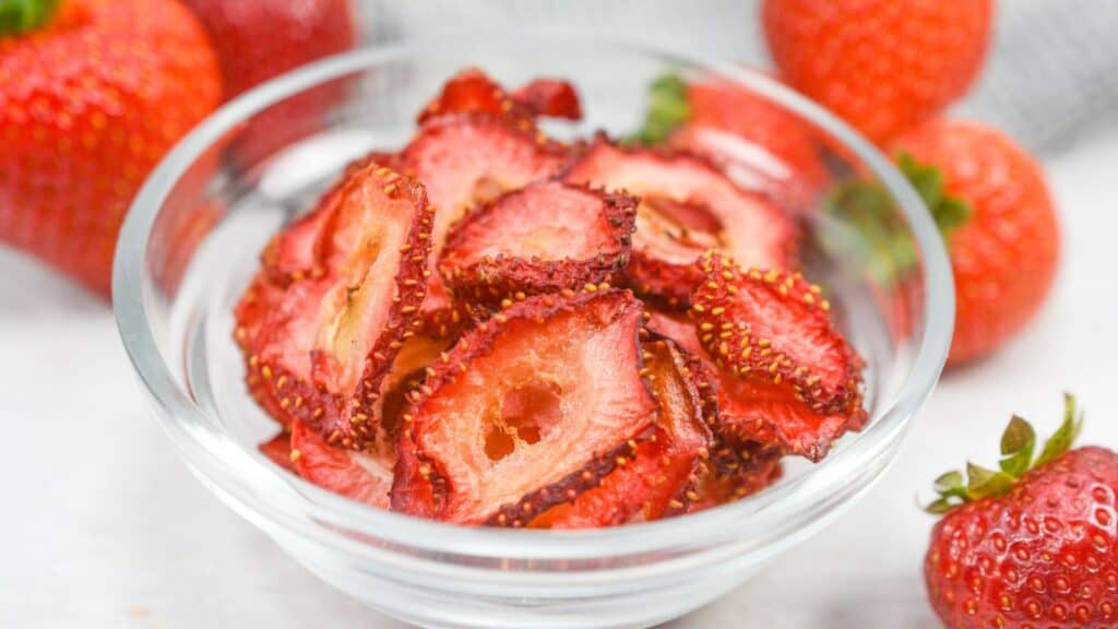 A clear bowl of dried strawberry slices on a white surface, with fresh whole strawberries in the background.