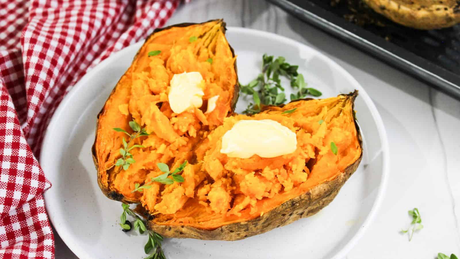 Two halves of baked sweet potato topped with butter and garnished with herbs on a white plate, with a red checkered napkin on the side.