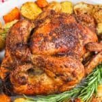 A roasted chicken surrounded by herbs and roasted potatoes and carrots on a white plate.