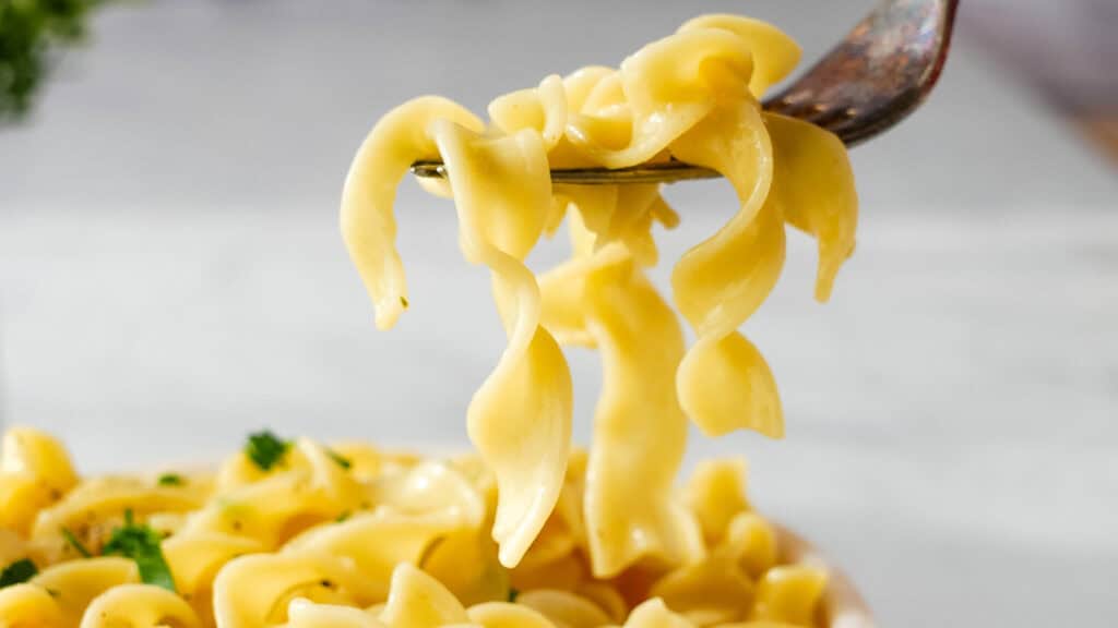 A close-up image of creamy fettuccine pasta being lifted with a fork, with more pasta and parsley visible in the background.