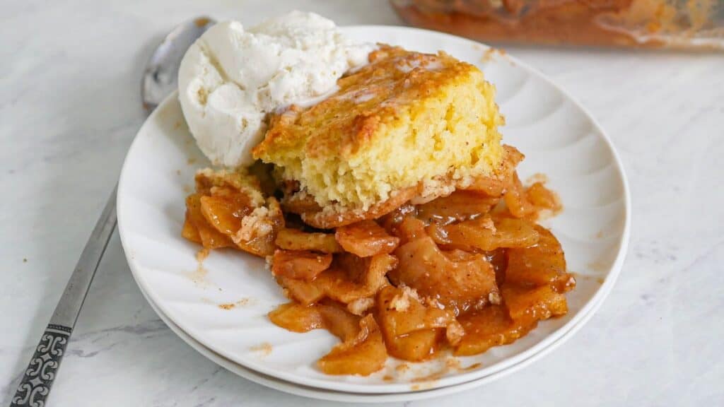 A slice of apple cobbler topped with a scoop of vanilla ice cream on a white plate, with a fork to the side.