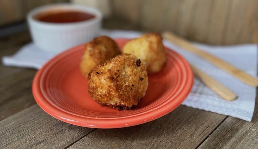 Three golden-brown arancini balls on an orange plate with a side of marinara sauce, on a rustic wooden table.