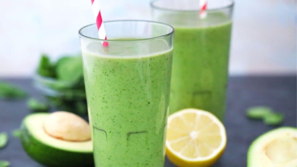 Two glasses of green smoothie with a lemon half and avocado in the background.