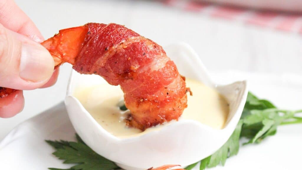 A hand dipping a bacon-wrapped shrimp into a small bowl of sauce, placed on a white surface with a green leaf garnish.