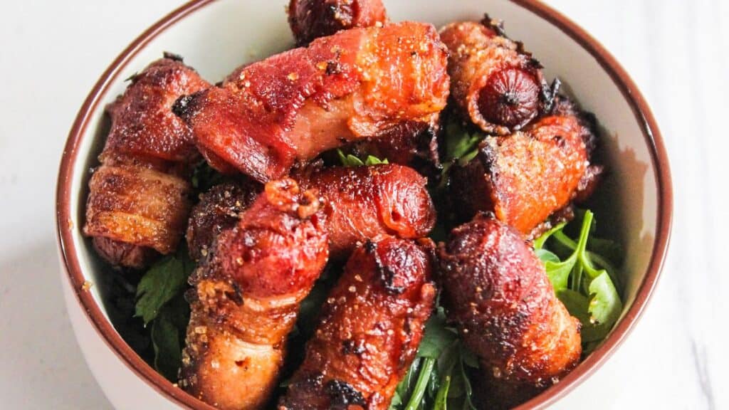 A bowl of bacon-wrapped cocktail sausages served on a bed of fresh green herbs.