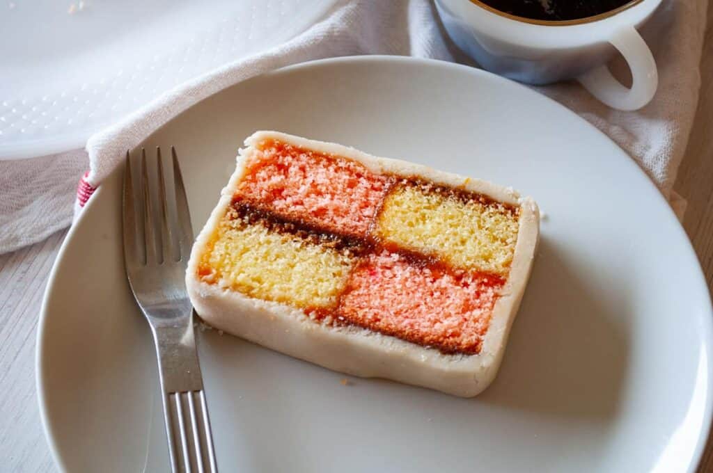 A slice of battenberg cake on a plate with a fork, accompanied by a cup of coffee.
