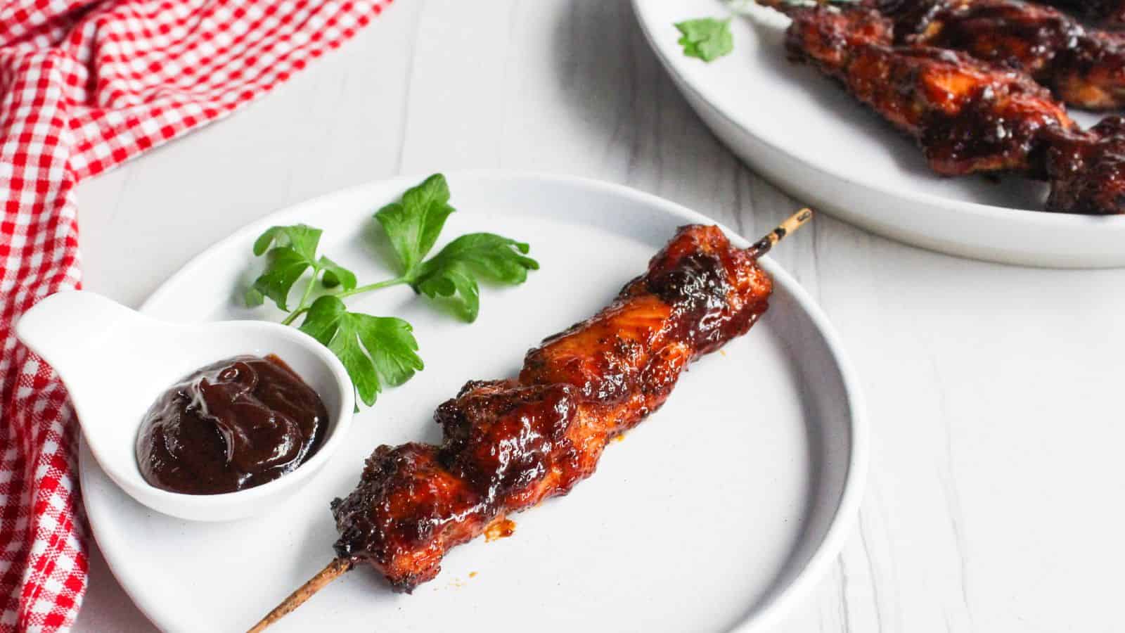 A single barbecued chicken skewer on a white plate, with a small bowl of barbecue sauce and a parsley garnish, next to a plate of additional chicken skewers.