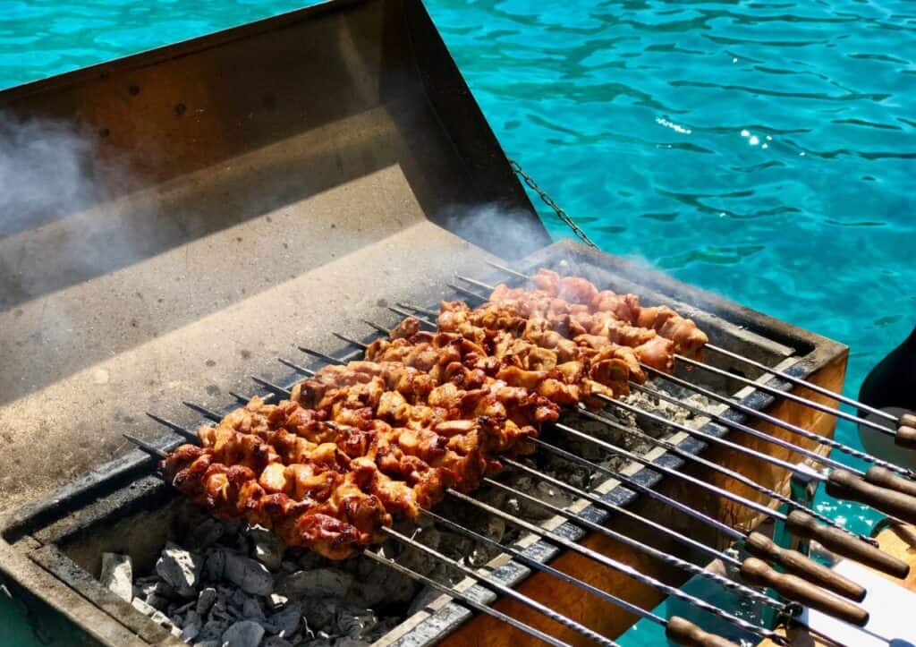 Barbecue skewers cooking over a charcoal grill on a boat with clear blue water in the background.