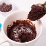 A spoon lifting a bite of freshly made chocolate mug cake from a white mug, with chocolate chips in a small bowl in the background.