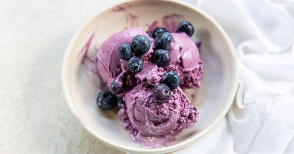 Bowl of blueberry ice cream topped with fresh blueberries on a light surface.