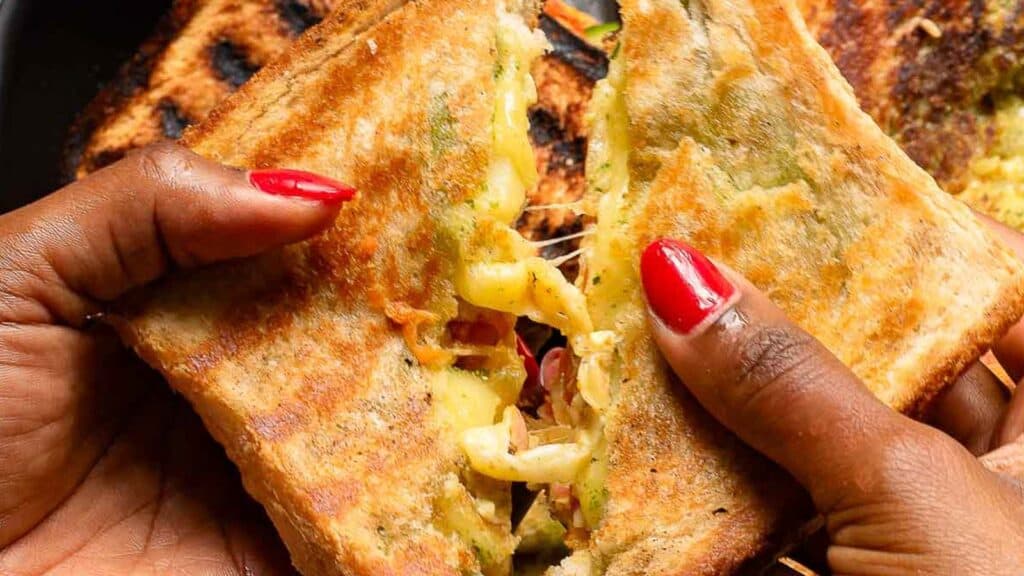 Hands with red nail polish holding a grilled cheese sandwich with visible melted cheese and fillings.