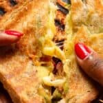 Hands with red nail polish holding a grilled cheese sandwich with visible melted cheese and fillings.