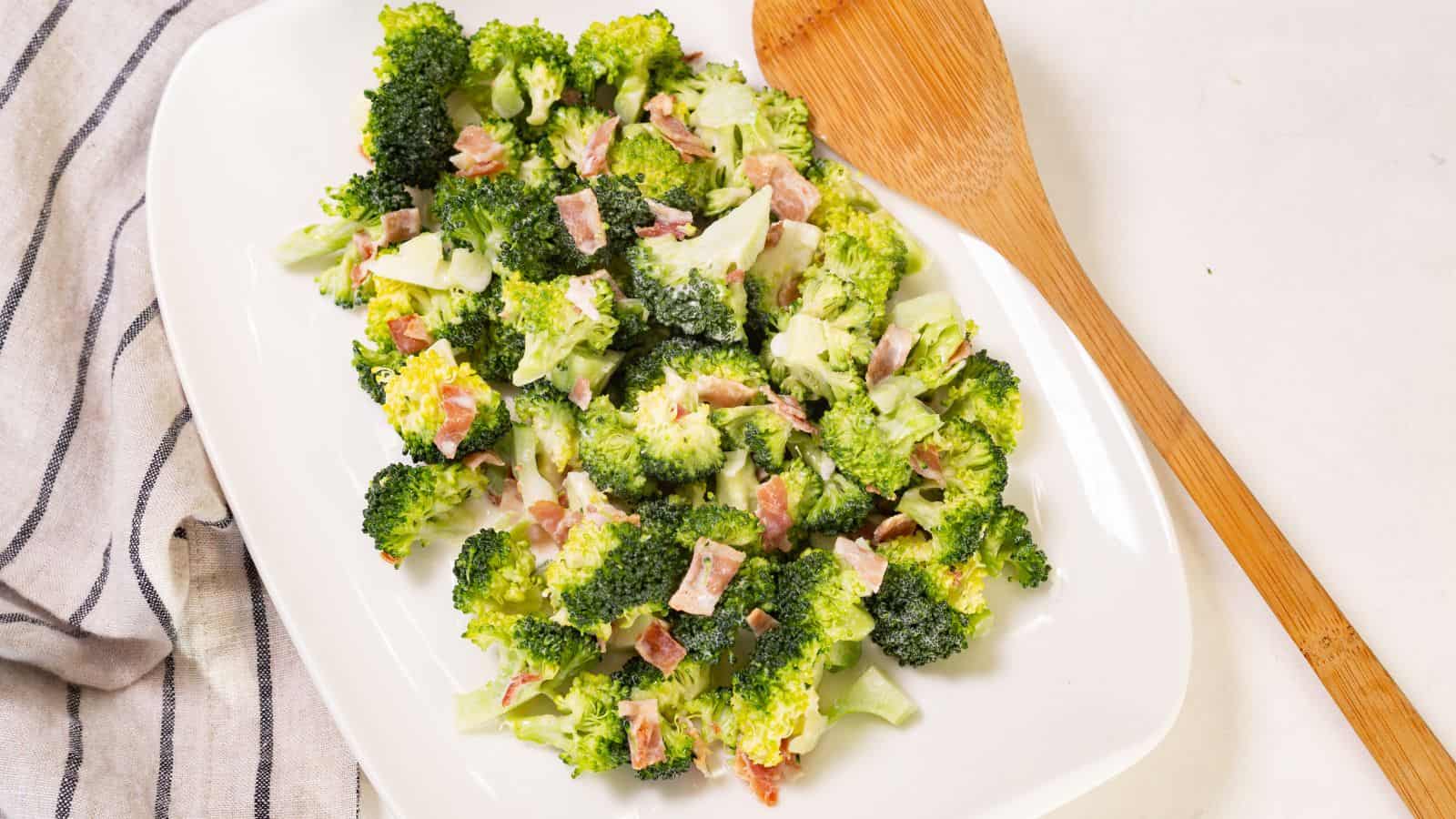 Broccoli salad with bacon bits on a white oval plate, accompanied by a wooden serving spoon on a linen napkin.