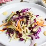 A plate of colorful broccoli slaw with shredded purple cabbage and carrots topped with dried cranberries and pine nuts, next to a red-checked napkin.