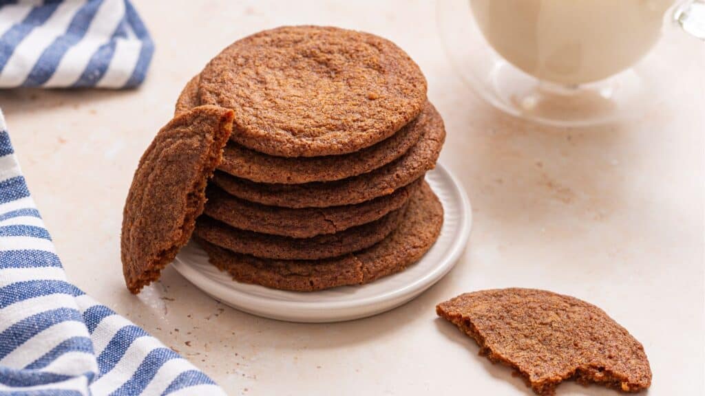 A stack of brown cookies on a small white plate with one cookie broken in half, next to a glass of milk and a striped blue and white napkin.