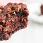 Close-up of a moist chocolate brownie with melted chocolate chunks, held between fingers with a blurred plate in the background.