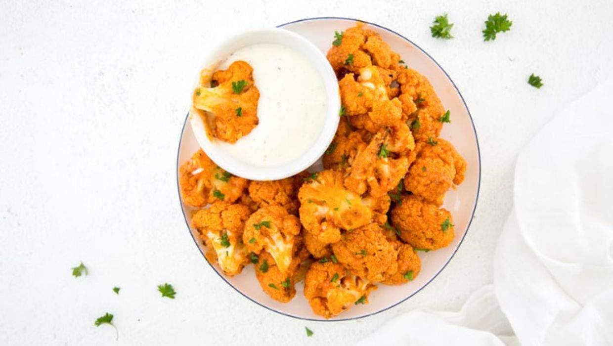 A plate of buffalo cauliflower bites served with a side of ranch dressing, garnished with parsley on a white surface.