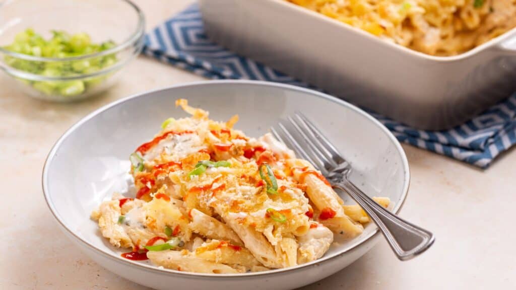 A plate of cheesy baked pasta and chicken casserole served with chopped scallions, accompanied by a baking dish and fork.