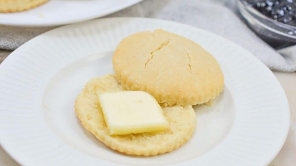 Biscuit cut in half on a white plate, one half with a pat of butter on top, next to a cloth napkin and a bowl of jam in the background.