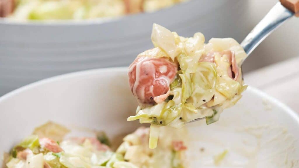 A fork lifting a serving of cabbage and sausage Alfredo from a bowl.